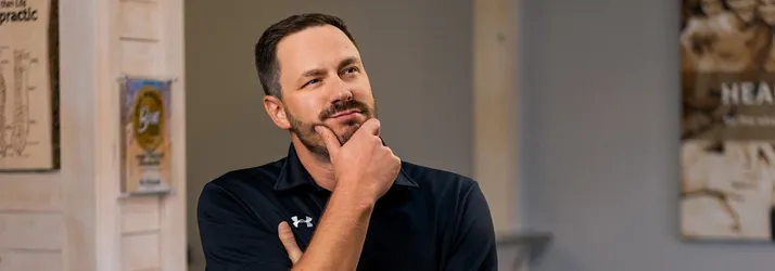 Dusty Large in a chiropractic clinic wearing a dark polo shirt and posing with a hand on the chin near a reception area.