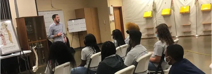 Dr. Dusty Large speaking to a seated audience in a classroom setting, with anatomical charts and presentation materials visible.
